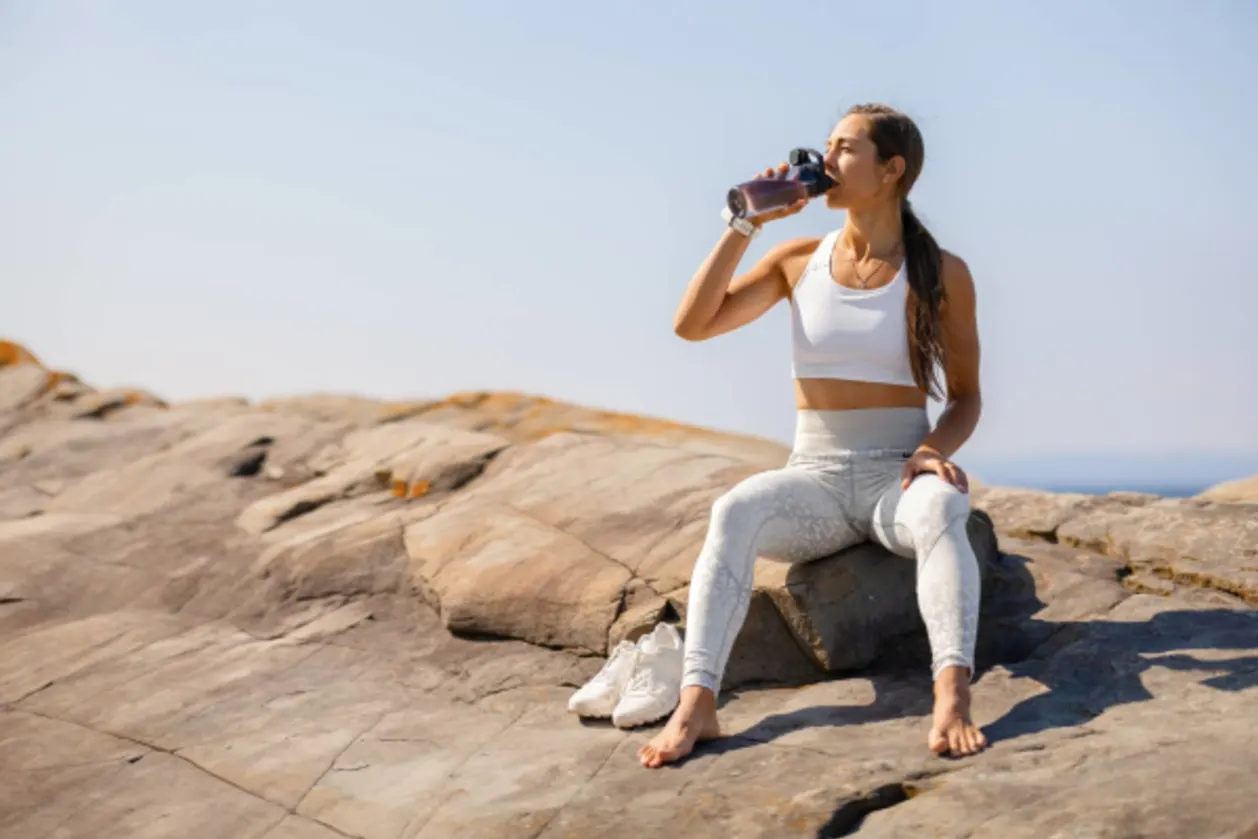 Fit woman hydrates by drinking water while sitting on a rocky coastal landscape